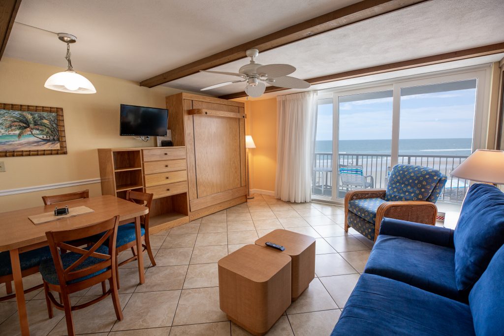 A wide view of a living room with a dining area, a sofa, and a balcony with an ocean view.