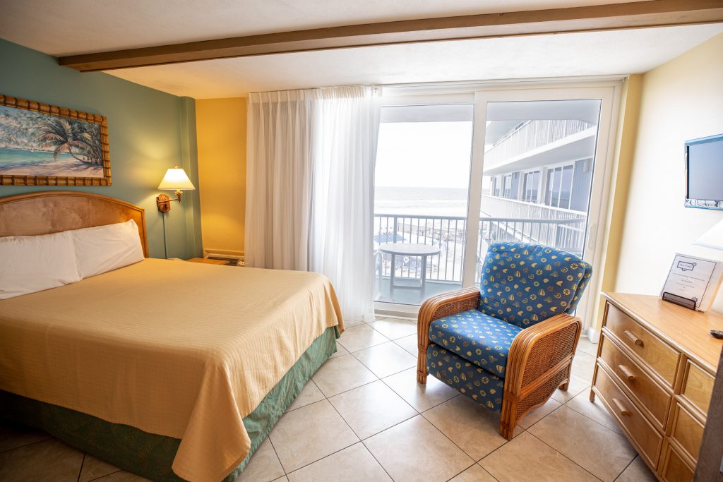 A wide view of a hotel room with a bed, chair, dresser, and balcony with an ocean view.