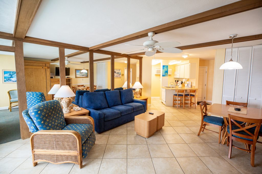 A wide view of a hotel living room with a blue sofa, wooden beams, a ceiling fan, and a kitchenette.